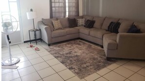 Ein Wohnzimmer mit einem beigefarbenen Ecksofa, einem kleinen Beistelltisch, einer Stehlampe und einem gemusterten Teppich.