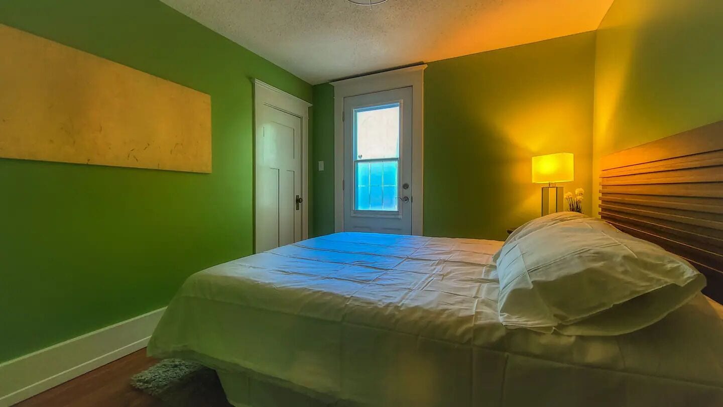 A bedroom with a bed, a nightstand with a lamp, a door, and a window.