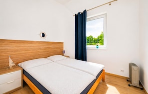 Un dormitorio con cabecera de madera, una cama con colchón blanco, una ventana con vistas a la vegetación, una mesita de noche con lámpara y un radiador.