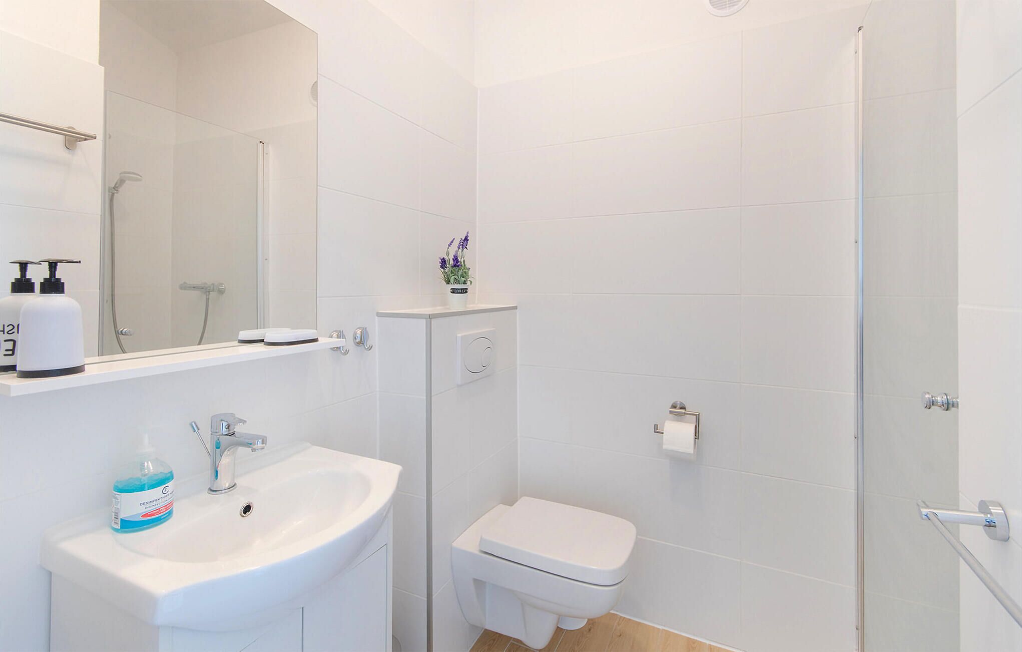 A modern bathroom with a white sink, toilet, and shower. There are toiletries on the sink and a small plant on the wall.