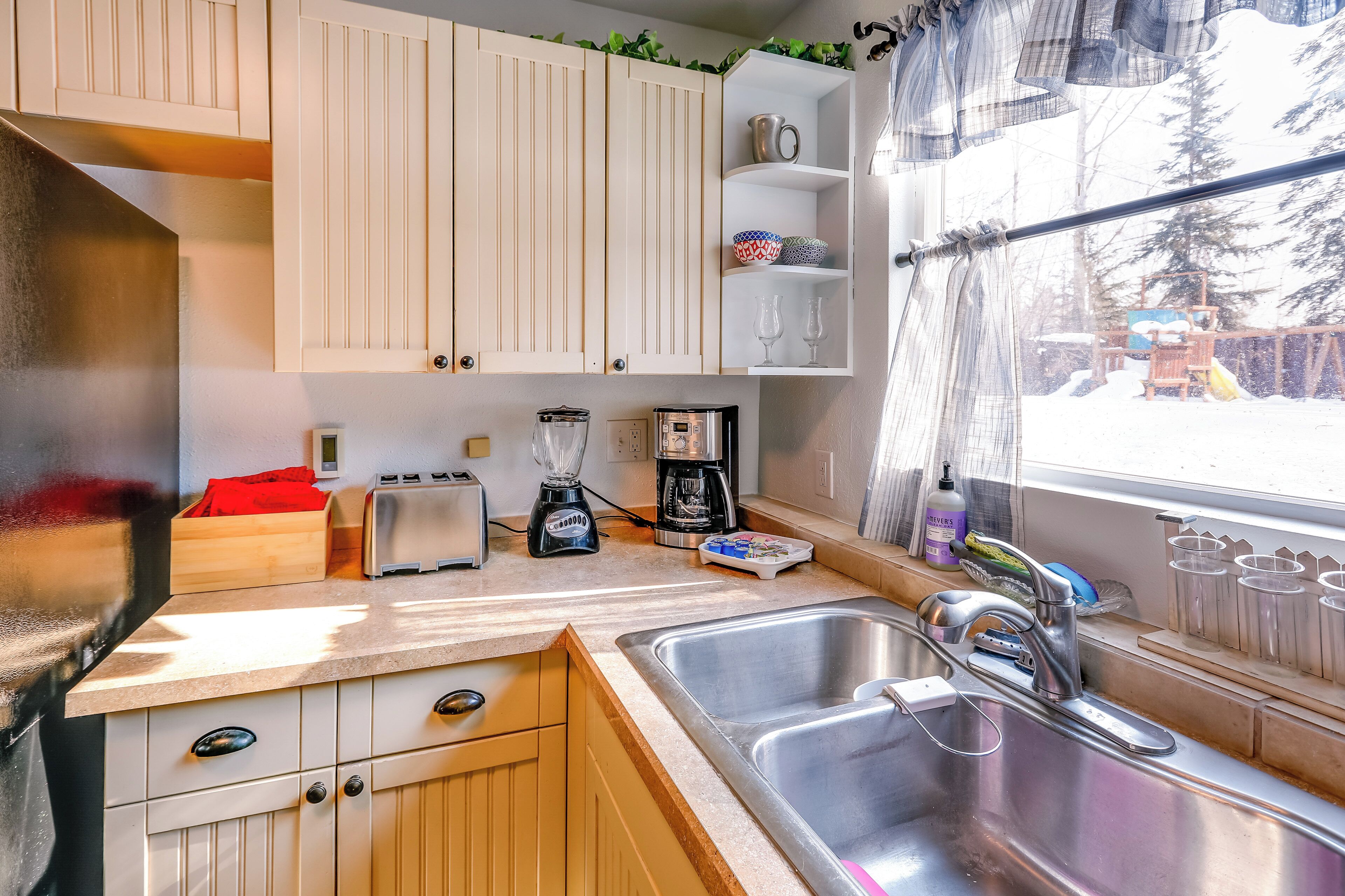 Een compacte keuken met witte kasten, een spoelbak, een broodrooster, een koffiezetapparaat en een raam met uitzicht op bomen.