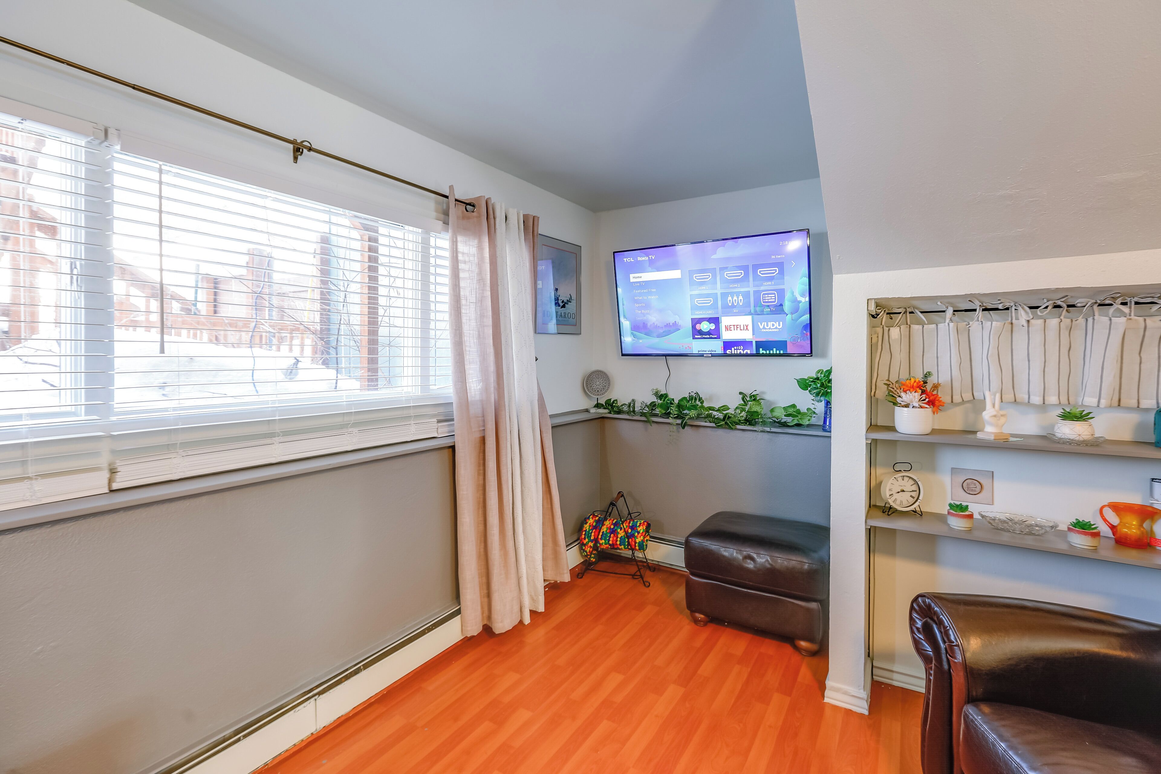 A living room with a wooden floor, a TV mounted on the wall, a cushioned bench, and a shelf with decorative items.