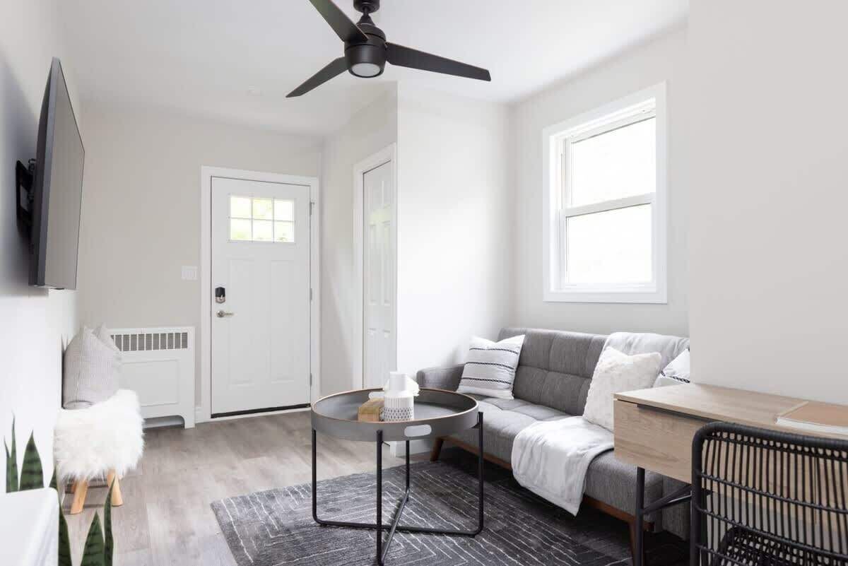 A modern living room with a grey sofa, a round coffee table, a ceiling fan, and a large window providing natural light.