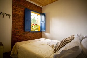 Cozy bedroom with a bed against a brick wall, featuring a window with blue shutters and simple, functional design.