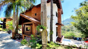 Uma cabana rústica com varanda de madeira, rodeada por vegetação exuberante e montanhas ao fundo.