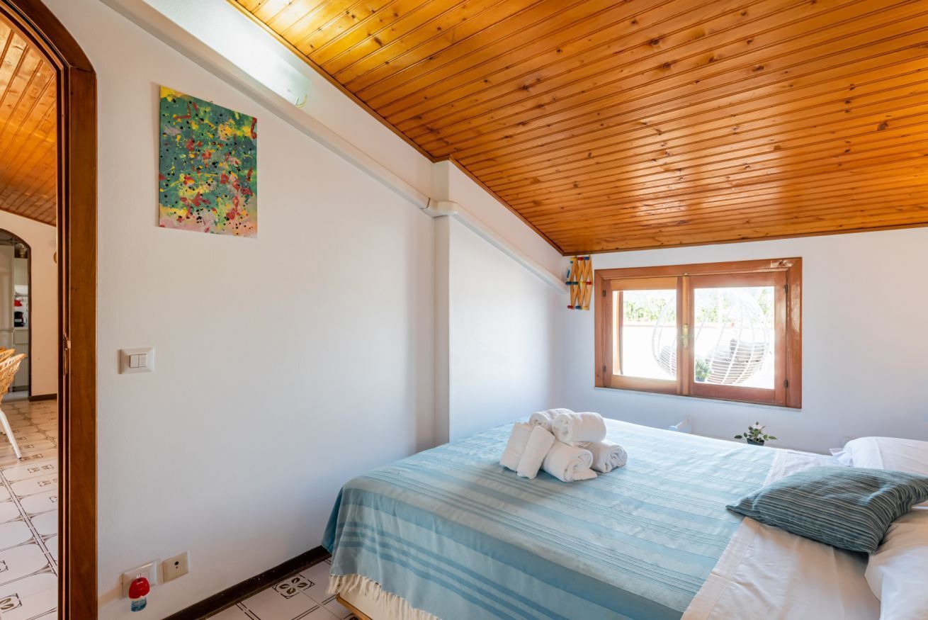A bedroom with a wooden ceiling, a bed with a blue and white striped bedspread, a window with a view of greenery, and a colorful abstract painting on the wall.