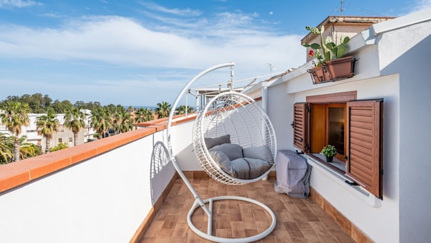 Un balcón con una silla colgante blanca, una ventana con contraventanas de madera y una planta en maceta.