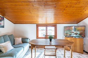 A room with a wooden ceiling, a dining table with chairs, a sofa, a TV, and a window with a view of a garden.