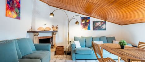 A cozy living room with a wooden ceiling, a fireplace, blue sofas, a wooden table, and artwork on the walls.