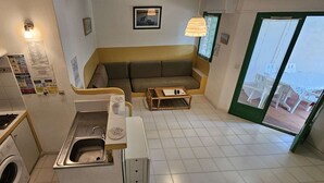 Une cuisine compacte équipée d’un four à micro-ondes, d’une cuisinière, d’un évier et d’un lave-linge. Le coin salon comprend un canapé et une petite table avec une lampe, et offre une vue sur l’extérieur.
