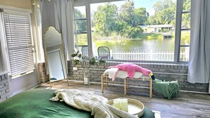Ein Schlafzimmer mit großem Fenster mit Blick auf einen See, ein Bett mit grüner Bettdecke, ein Sessel mit rosa Decke und ein kleiner Tisch mit Kerzen.