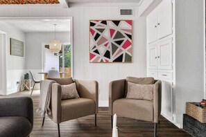 Ein modernes Wohnzimmer mit zwei beigen Sesseln, einer weißen Pendelleuchte und einem geometrischen Wandbild.