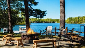 Un espacio al aire libre junto al lago con una fogata, bancos y sillones de descanso.