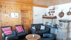 A cozy room with wooden walls and a stone pillar. There are three black chairs with patterned cushions, a round wooden table, and a shelf with kitchen utensils. - Outside: Chalet of about 132m2, for 10 people, with garden and garden furniture -Inside: living room (Chandolin)