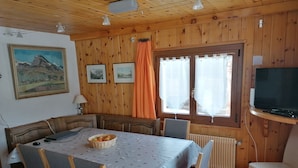 A dining area with a table set for two, a television, and a window with curtains. - Outside: Chalet of about 132m2, for 10 people, with garden and garden furniture -Inside: living room (Chandolin)