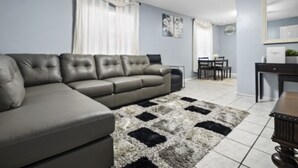 Un salon comprenant un canapé d’angle, un ventilateur de plafond, un coin repas avec une table et des chaises, et un tapis à motifs noir et blanc.