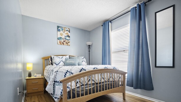 Une chambre à coucher avec un lit en bois, une table de chevet avec une lampe, une œuvre d’art accrochée au mur et une fenêtre avec des rideaux.