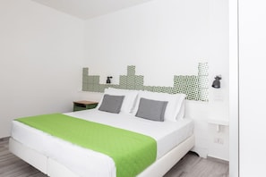 Una habitación de hotel con una cama, una manta verde, ropa de cama blanca, una mesita de noche, una lámpara y una pared con baldosas de diseño geométrico.