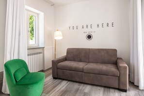 Una habitación con un sofá marrón, una silla verde, una ventana con cortinas blancas y una pared con texto motivacional.