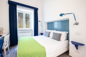 A hotel room with a bed, a desk, a chair, a window with curtains, a blue wall lamp, and a blue wall shelf.