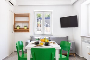 A dining area with a white table, green chairs, a grey sofa, a TV mounted on the wall, and a window with a view of the outside.