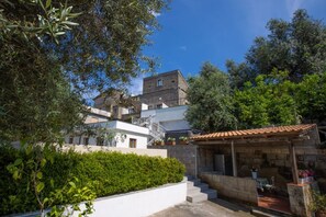 A hillside villa with a stone terrace, a white building with a balcony, and a garden area.