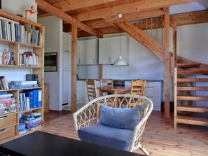 Ein gemütliches Zimmer mit Holzboden, ein Korbstuhl mit Kissen, ein Esstisch und ein Bücherregal voller Bücher und Deko-Artikel.