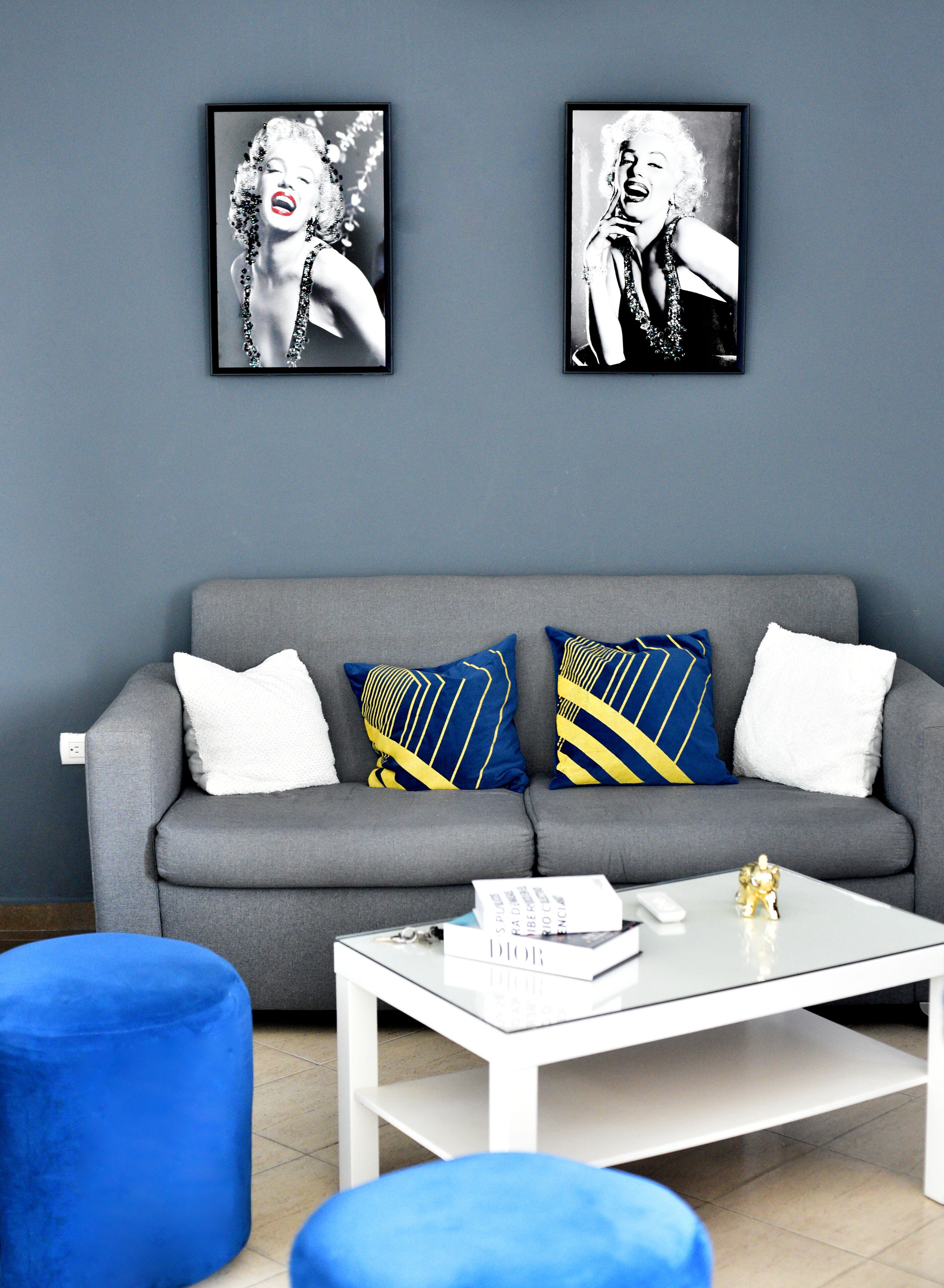 A living room with a grey sofa, blue and yellow patterned cushions, a white coffee table with a book and remote control, and two framed pictures on the wall.