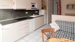 Una cocina compacta con armarios blancos, un microondas y un lavamanos. Hay un comedor con una mesa y una silla, y un sofá a cuadros en la sala de estar.