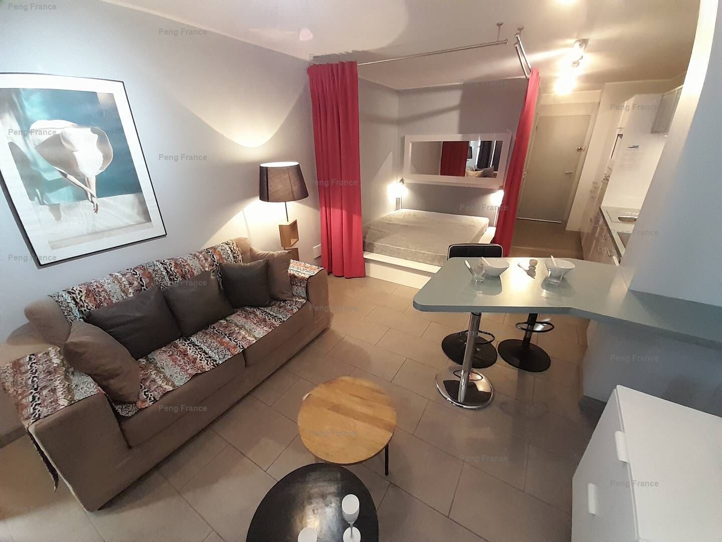 A modern living room with a sofa, a coffee table, a dining table with chairs, a red curtain, and a bed in the background.