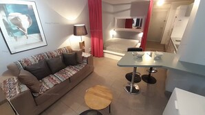 A modern living room with a sofa, a coffee table, a dining table with chairs, a red curtain, and a bed in the background. - Apartement Intense (LE CAP D AGDE)