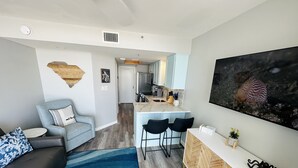 Un soggiorno moderno con un divano grigio, un cuscino azzurro decorato, un tavolino da caffè in marmo e un tappeto blu. È presente un angolo cottura con un piano di lavoro bianco e una piccola isola con due sgabelli neri. Al muro, un grande quadro raffigura una conchiglia.