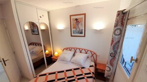 A bedroom with a bed, bedside lamps, a mirror, a framed picture on the wall, and a window with curtains. - Villa COCO GIRL (LE CAP D AGDE)