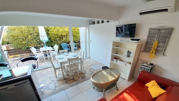 A living room with a red sofa, a TV on a white shelf, and a dining area with white chairs and a table. - Villa Tabou (LE CAP D AGDE)