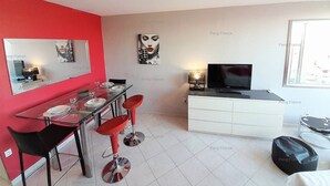 A modern living room with a red accent wall, a dining area with a glass table and red bar stools, a white entertainment unit with a TV, and a window with a view. - Apartment "PEPPERONI" (LE CAP D AGDE)