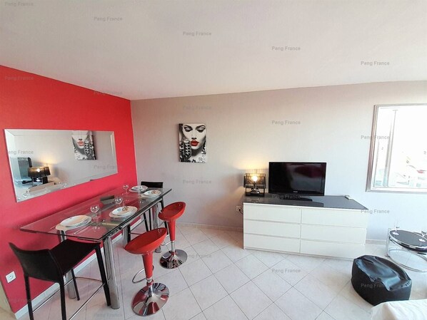 A modern living room with a red accent wall, a dining area with a glass table and red bar stools, a white entertainment unit with a TV, and a window with a view.