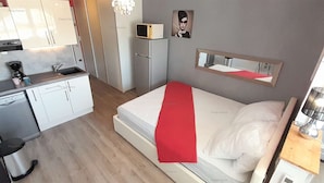 Ett kompakt hotellrum med ett litet pentry, en säng med ett rött överkast, ett sängbord med en lampa och en spegel på väggen.