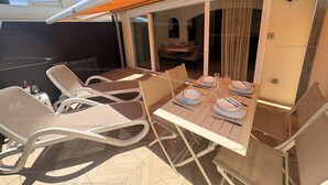 An outdoor dining area with a table set for two, cushioned chairs, and a sliding glass door leading to an interior space.