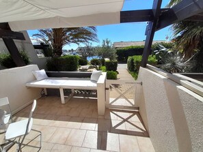 An outdoor patio area with a white sofa, a small table, and a chair. There is a white umbrella providing shade. The patio is surrounded by greenery and a view of the street.
