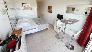 A compact hotel room with a bed, a small desk with a chair, a mirror, and an air conditioning unit. - Studio Piña Colada (LE CAP D AGDE)