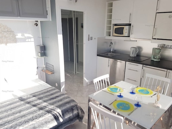 A compact kitchen with a microwave, coffee maker, and a sink. There is a dining table with a white chair and a bed with a gray blanket.