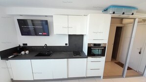 A modern kitchen with white cabinets, a black countertop, a built-in oven, and a flat-screen TV mounted above the sink. - Studio Ô Plaisir (LE CAP D AGDE)