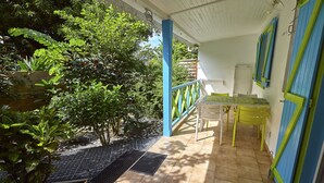 Une terrasse couverte avec une table et des chaises, entourée de verdure et dotée d’un sol carrelé.