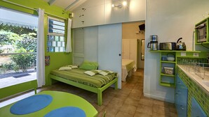 Une chambre de taille modeste comprenant un lit, une petite table, un coin cuisine et donnant sur un jardin.