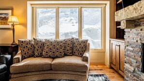 A cozy living room with a brown sofa, patterned cushions, a fireplace, and a view of a snowy landscape through a large window. - Beautiful Modern Home in Great Location, Hot Tub +Patio, Luxury Amenities! (Park City)