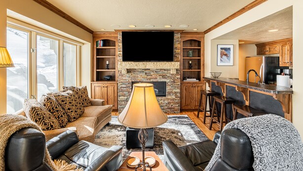 A cozy living room with a fireplace, a television, and a kitchen area with wooden cabinets and a bar. - Beautiful Modern Home in Great Location, Hot Tub +Patio, Luxury Amenities! (Park City)