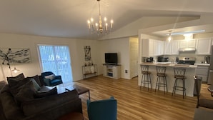 Een moderne woonkamer met een bank, een televisie en een kroonluchter. Er is een keuken met witte kasten en een bar.