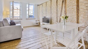 Ein modernes Wohnzimmer mit einem weißen Sofa, einem Esstisch und einem Fernseher. Fenster bieten einen Blick auf die Gebäude draußen.