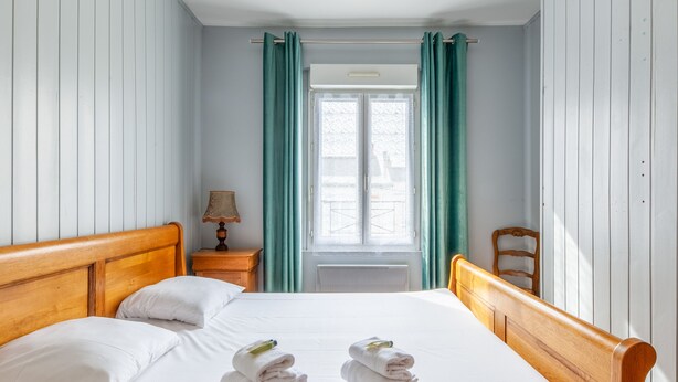 A neatly made bed with white linens and two towels, a wooden headboard, a nightstand with a lamp, and a window with green curtains. - Maison Flatelle - Welkeys (Trouville-sur-Mer)