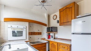 A kitchen with a built-in oven, microwave, and refrigerator. There is a sink with a faucet, wooden cabinets, and a clock on the wall. - Maison Flatelle - Welkeys (Trouville-sur-Mer)